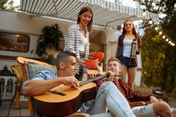 Happy friends sing songs with guitar on picnic at camping in the forest. Youth having summer adventure on rv, camping-car on background. Two couples leisures, travelling with trailer