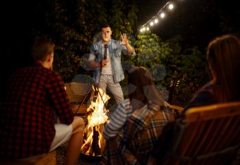 Friends tell scary stories by the bonfire, picnic at camping in the forest. Youth having summer adventure on rv, camping-car on background. Two couples leisures, travelling with trailer