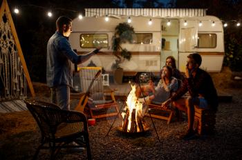 Man tells a scary story to his friends by the bonfire, picnic at camping in the forest. Youth having summer adventure on rv, camping-car on background. Two couples leisures, travelling with trailer