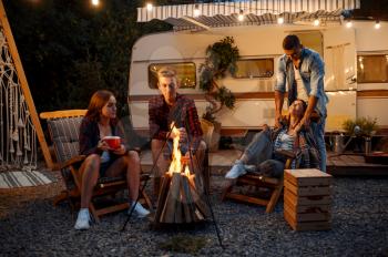 Friends warming themselves by the bonfire, picnic at camping in the forest. Youth having summer adventure on rv, camping-car on background. Two couples leisures, travelling with trailer