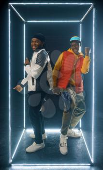 Two stylish rappers poses in glowing cube, studio with dark background. Hip-hop performers, trendy rap singers, break-dancers