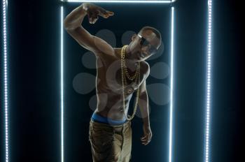 Serious rapper in gold chains dancing in illuminated cube, dark background. Hip-hop performer, rap singer, break-dance performing, entertainment lifestyle, breakdancer