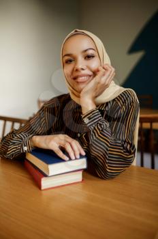 Arab female student in hijab holds textbook in university cafe. Muslim woman with books sitting in library