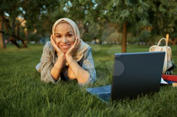 Arab female student in hijab lying on the grass with laptop in summer park. Muslim woman with books resting on the lawn. Religion and education