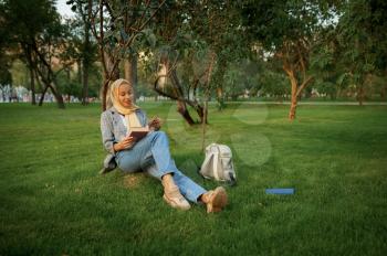 Arab female student in hijab reading textbook in summer park. Muslim woman with books resting on the lawn. Religion and education