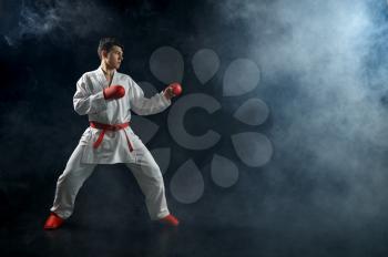 Male karate fighter in white kimono and red gloves, combat stance, dark background. Fighter on workout, martial arts, training before fighting competition