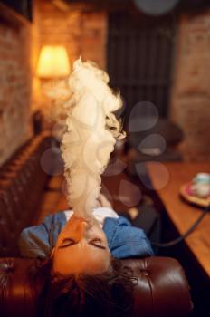 Young woman smokes on sofa in hookah bar, mesmerizing cloud. Shisha smoking, traditional bong culture, tobacco aroma for relaxation, rest with hooka