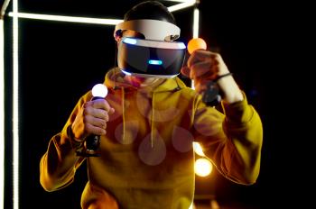 Young gamer plays in virtual reality headset and gamepad in luminous cube, front view. Dark playing club interior, spotlight on background, VR technology with 3D vision