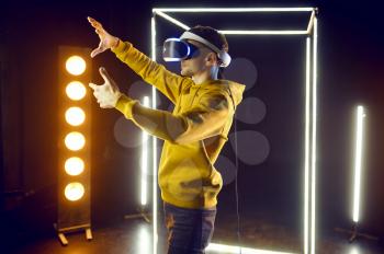 Young gamer plays the game using virtual reality headset and gamepad in luminous cube, front view. Dark playing club interior, spotlight on background, VR technology with 3D vision