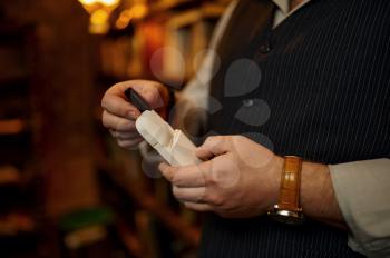 Man holds a pack of cigarettes and mouthpiece, bookshelf and rich office interior on background. Tobacco smoking culture, specific flavor. Smoke habit