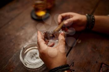 Man with guillotine cuts a cigar, wooden table on background. Tobacco smoking culture, specific flavor. Male smoker leisures with glass of alcohol