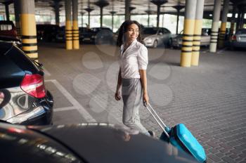 Young woman with suitcase in car parking. Female traveler with luggage in vehicle park lot, passenger with bag. Girl with baggage near automobile