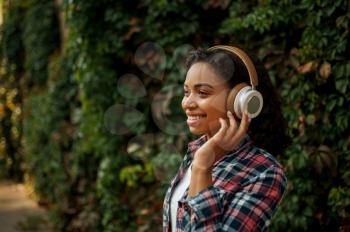 Happy woman in headphones listening to music in summer park. Female music fan walking outdoors, girl in earphones, outdoors