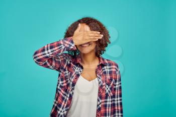 Cute young woman covers her eyes with hand, blue background, positive emotion. Face expression, female person looking on camera in studio, emotional concept, feelings