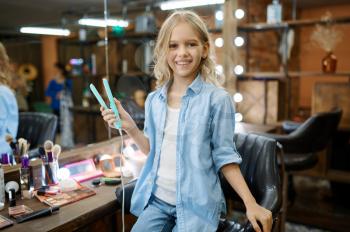 Female child holds hair straighteners in salon. Mom and little girl play hairdressers together, happy childhood, glamour family