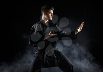 Male karate fighter in black kimono, combat stance, dark background. Man on workout, martial arts, fighting competition