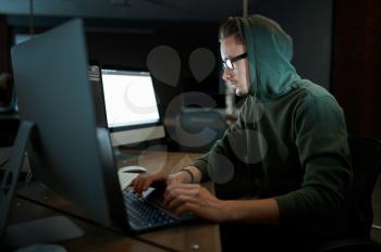 Male internet hacker in hood and glasses sitting at monitors. Illegal web programmer at workplace, criminal occupation. Data hacking, cyber security