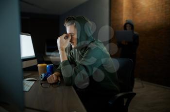 Tired male internet hacker in hood sitting at monitors. Illegal web programmer at workplace, criminal occupation. Data hacking, cyber security