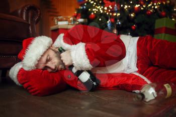 Vile drunk Santa claus sleeps under christmas tree with bottle of alcohol, nasty party, humor. Unhealthy lifestyle, bearded man in holiday costume, new year and alcoholism