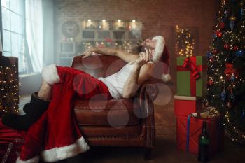 Vile Santa claus in red cap smoking cigar on couch, nasty party, humor. Unhealthy lifestyle, bearded man in holiday costume, new year and alcoholism