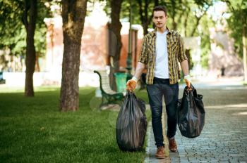 Young man holds plastic trash bags in park, volunteering. Male person cleans forest, ecological restoration, eco lifestyle, garbage collection and recycling, ecology care, environment cleaning