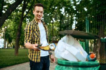 Young volunteer puts trash in plastic bin in park, volunteering. Male person cleans forest, ecological restoration, eco lifestyle, garbage collection and recycling, ecology care, environment cleaning
