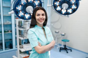 Smiling female surgeon in operating room, lamp and surgical table on background, surgery. Doctor in uniform, medical clinic worker, medicine and health, professional healthcare in hospital