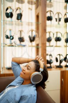 Happy woman listening to music in headphones store. Female person in audio shop, showcase with earphones on background, buyer in multimedia salon
