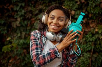 Music lover in headphones listening to music in summer park. Female audiophile walking outdoors, girl in earphones, green bushes on background