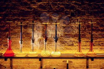 A row of hookahs with glass bulbs at the brick wall, nobody. Shisha bar equipment, traditional smoke culture, tobacco aroma for relaxation