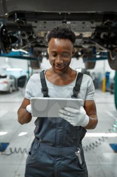 Male worker using laptop, car on lift, auto service. Vehicle repairing garage, man in uniform, automobile station interior on background