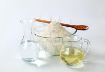 white wheat flour in a glass bowl with a wooden spoon, a carafe of cold water and a jug of sunflower oil 