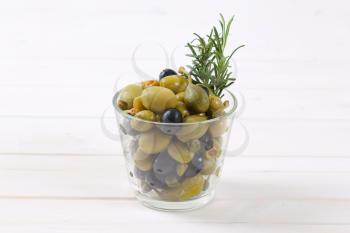 glass of pickled olives, capers and caper berries on white wooden background