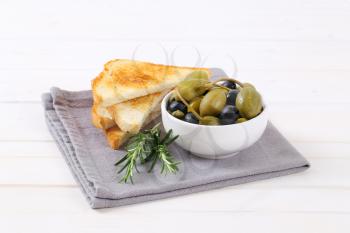 bowl of pickled olives, capers and caper berries with toast on grey place mat