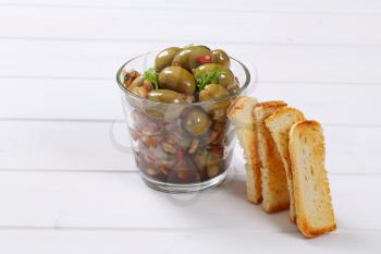 glass of marinated green olives with toast on white wooden background
