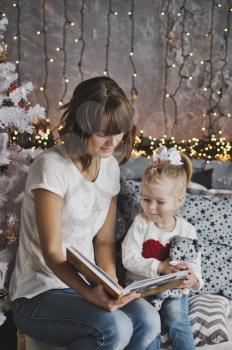 Little girl with mother reading a book sitting under the Christmas tree.