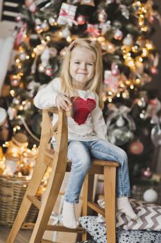 The child sits on a wooden chair beside the Christmas tree.