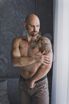 Portrait of a young bodybuilder with tattoos standing by the window.