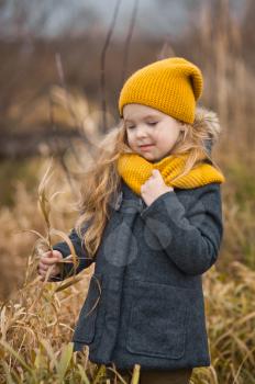 Little girl in a yellow hat and scarf walks along the shore of a river in autumn.
