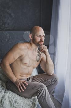 Bald brutal man sits pensively at the window.