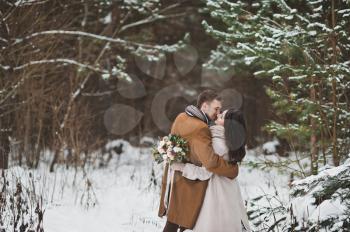 A gentle kiss of the newlyweds in the winter among the dark pine forest.