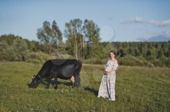 Portrait of a girl in a closed dress standing near a cow in a meadow.