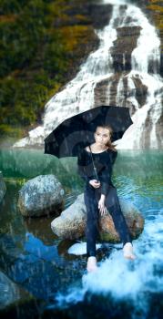 girl with an umbrella in the rain sitting the waterfall. She lowered her feet in the water.