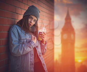 girl in a jeans jacket drinks juice on a Big Ben background in London