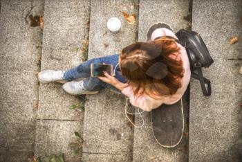 girl is sitting on a skateboard in headphones with a glass of coffee and a smartphone. View from above