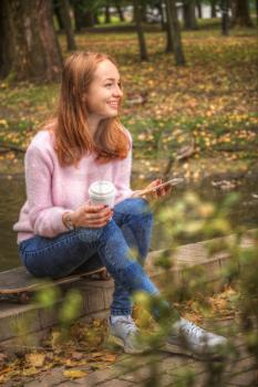girl is sitting on a skateboard in headphones with a glass of coffee and a smartphone. 