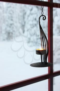 Decorative candle holder with burning candle hanging on window to the snowy winter yard with trees covered in snow