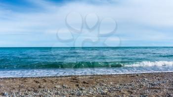 Blue sea with rocky pebble beach and waves with sea foam under blue sky with clouds in sunny day, beautiful nature landscape with horizon line