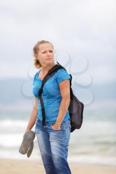 Pretty young blonde woman in a blue t-shirt and jeans with a backpack standing on a sea background.  Shallow depth of field. Focus on model. Vertical shot.