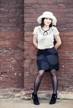Pretty young woman in a white hat, blouse and black skirt, posing outdoor against brick wall background. Toned photo with copy space. Vintage style photo.